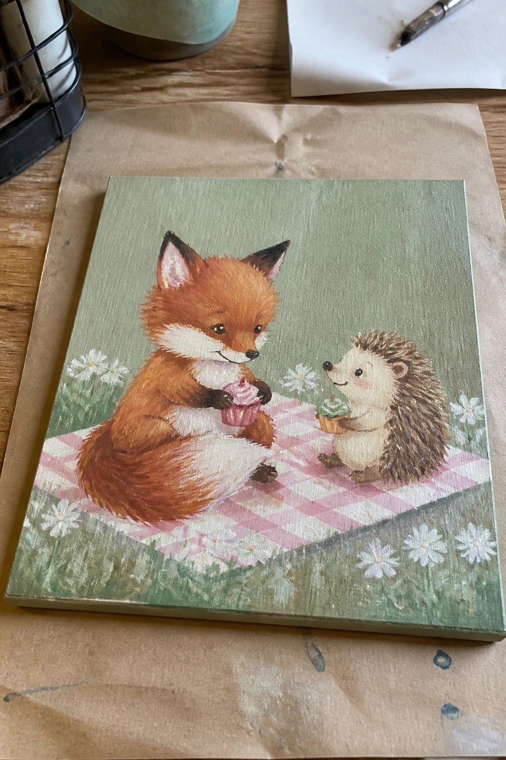 Charming painting of fox and hedgehog with cupcakes on pink checkered picnic blanket amid daisies