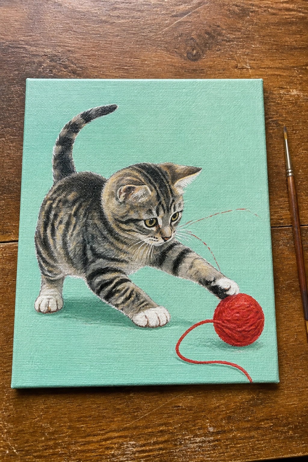 Realistic painting of tabby kitten reaching for red yarn ball on turquoise canvas.