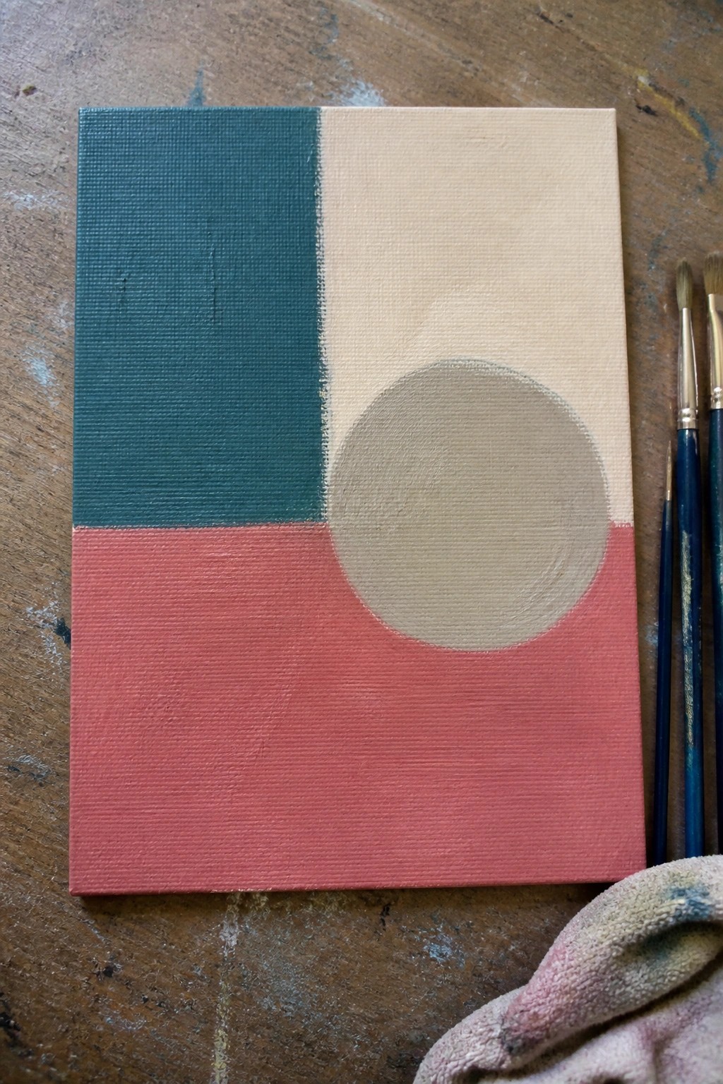Abstract painting with teal beige and pink sections plus gray circle beside paintbrushes