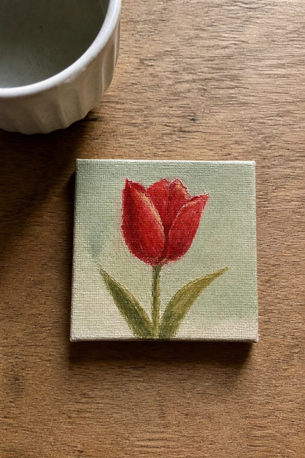 Miniature red tulip painting on canvas atop wooden table beside white bowl