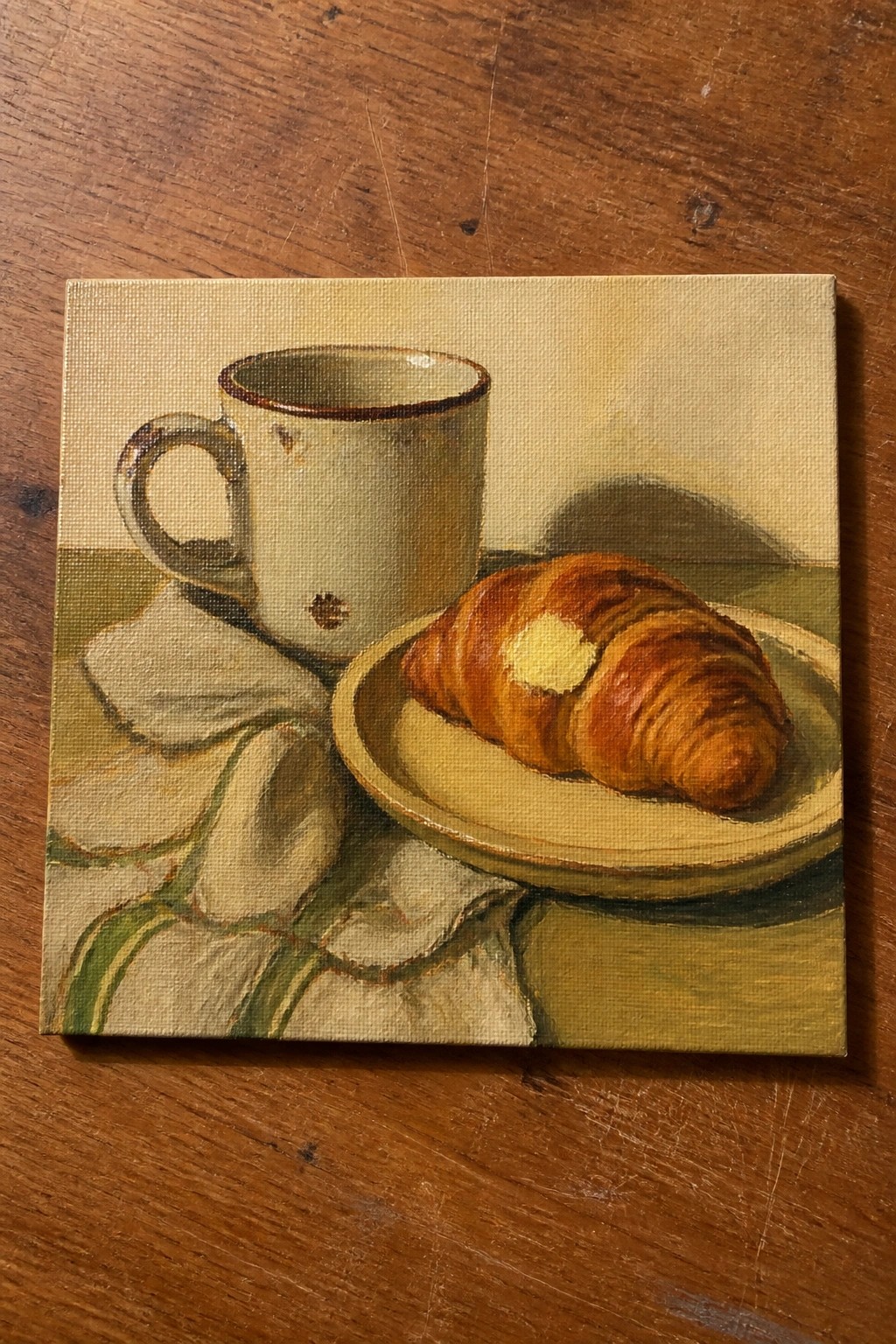 Still-life painting of buttered croissant on plate beside ceramic mug and striped napkin.