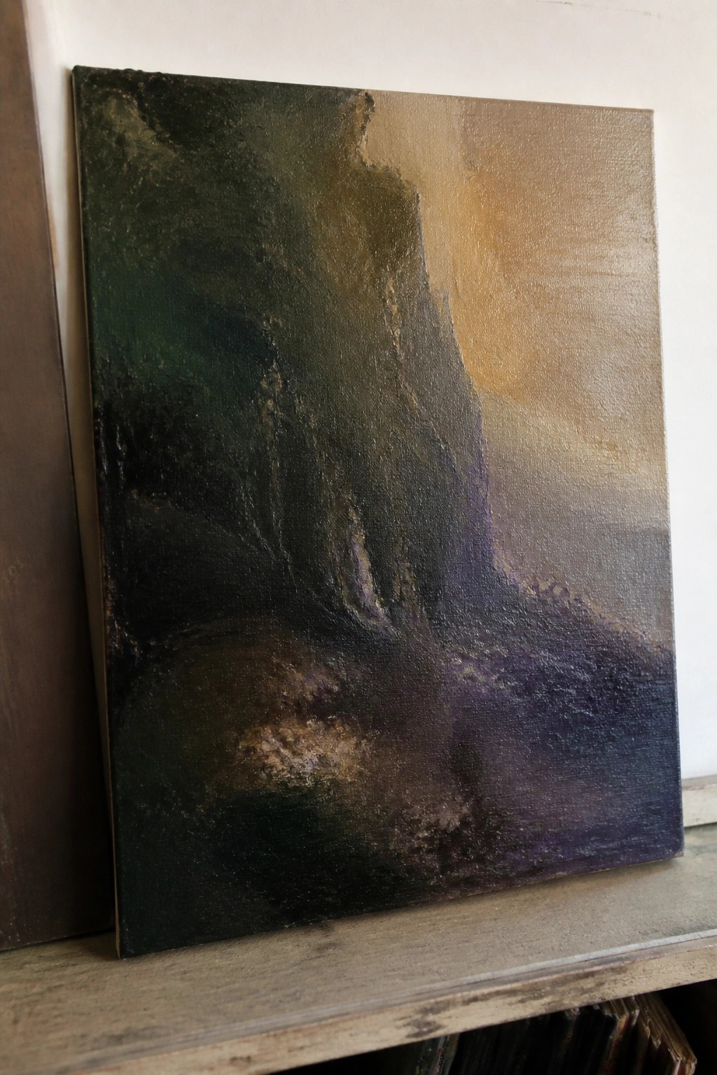 Abstract painting with textured dark greens, purples, and golden light on wooden shelf