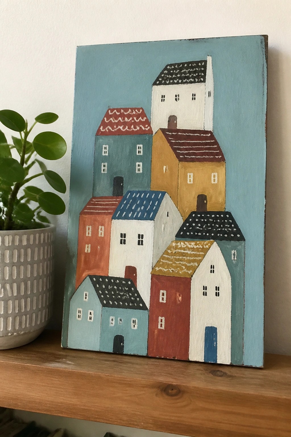 Colorful abstract houses painting on blue background beside potted green plant on shelf.