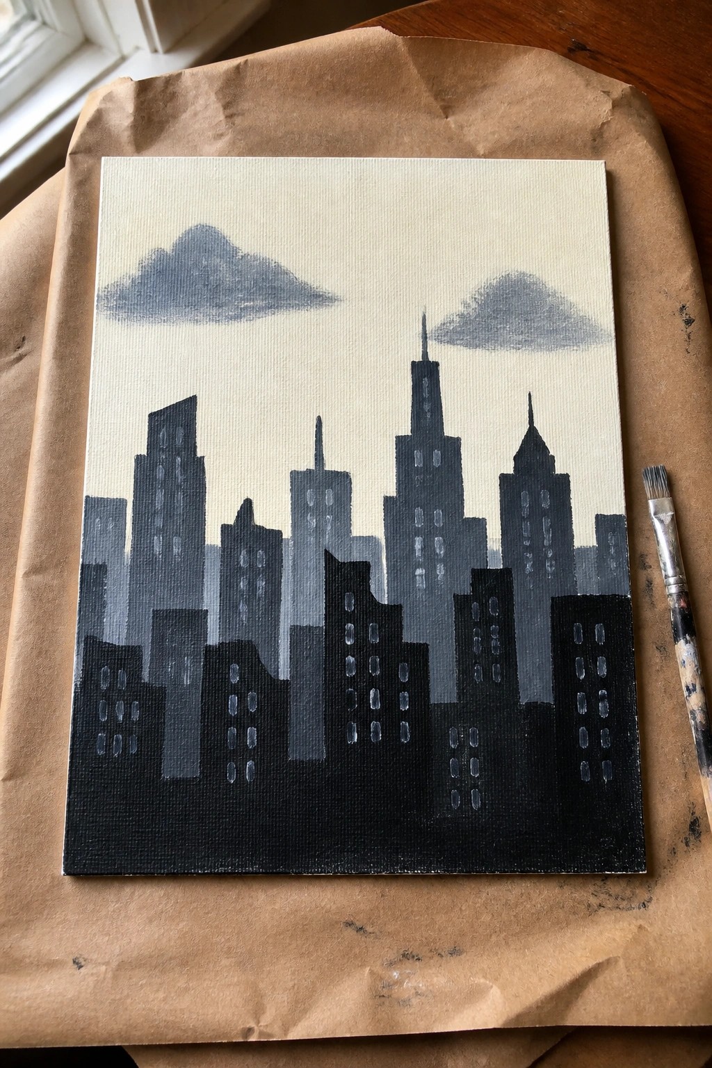Cityscape painting on canvas with dark buildings, gray clouds, and paintbrush on paper.