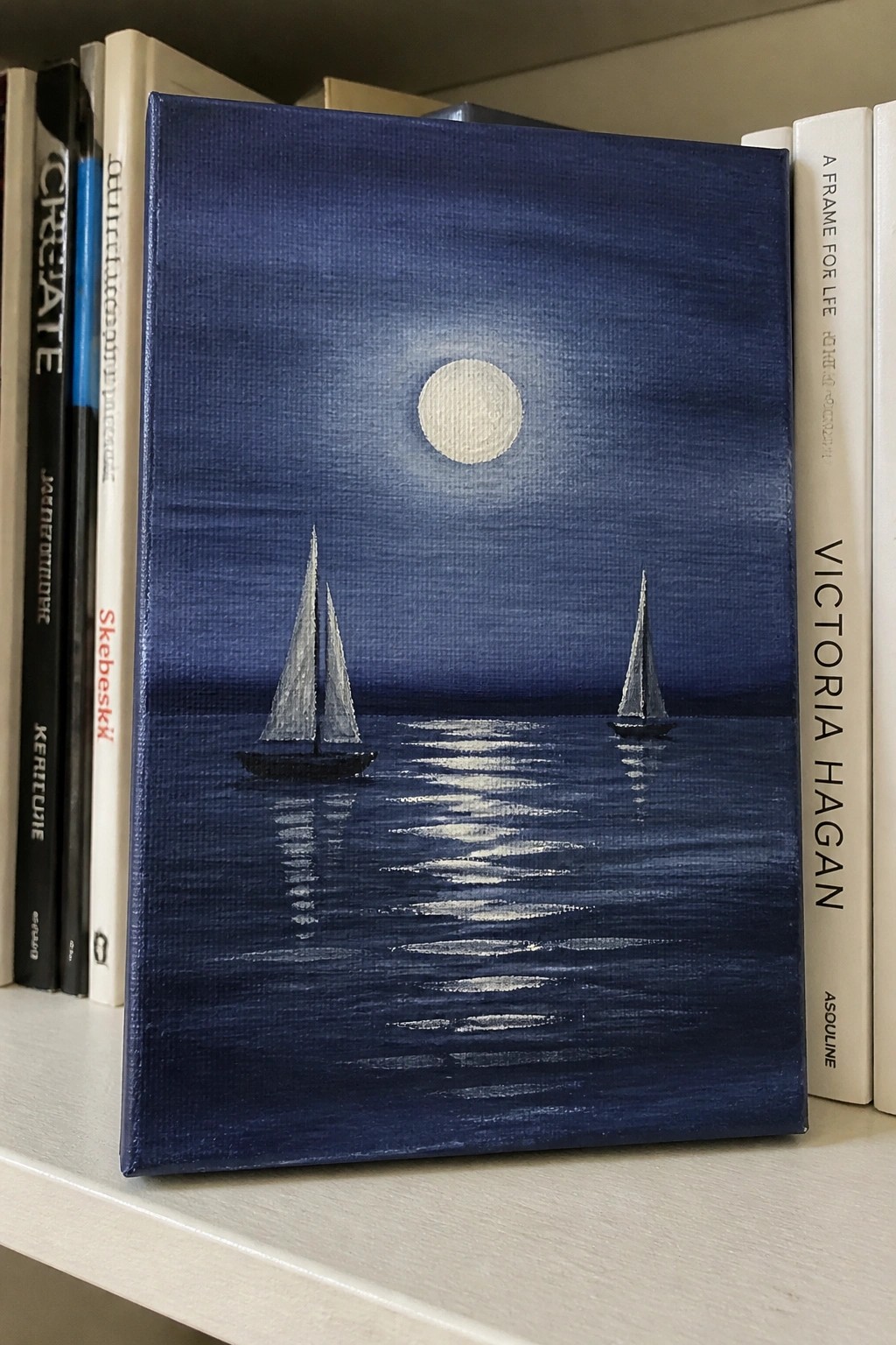 Canvas painting of two sailboats on moonlit sea between books on shelf