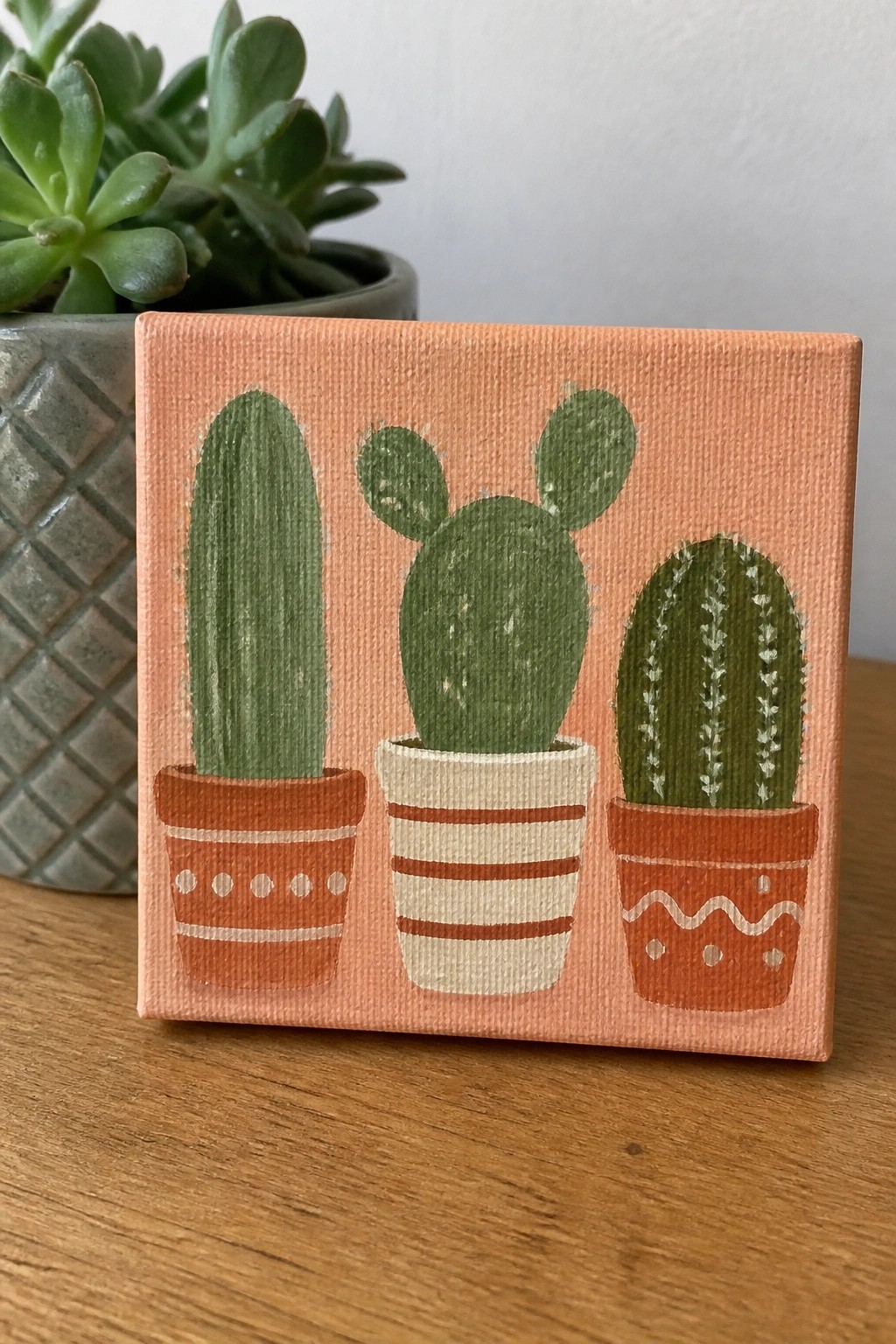 Three green cacti in patterned pots on a peach square canvas