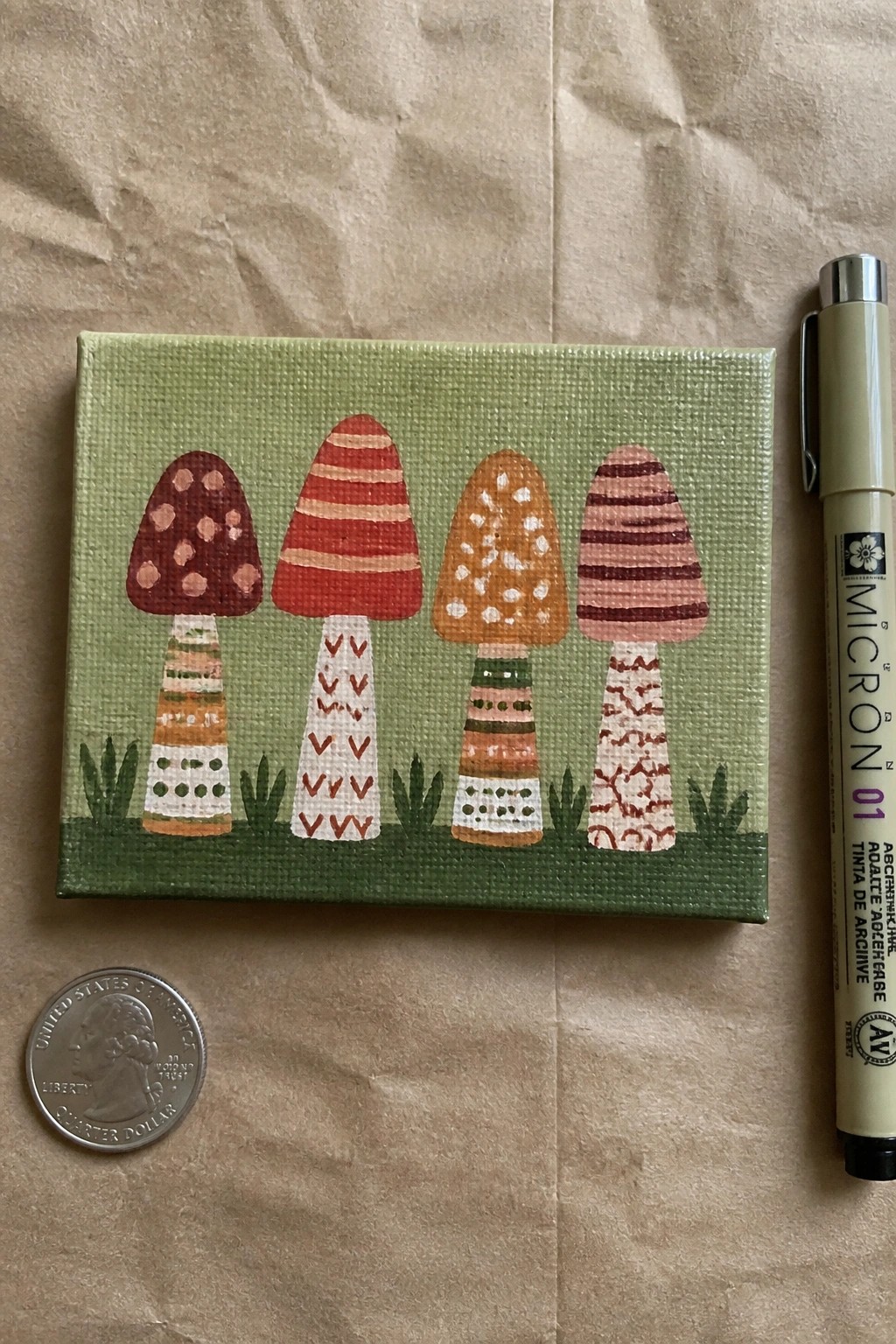 Small canvas painting of four colorful patterned mushrooms beside pen and quarter