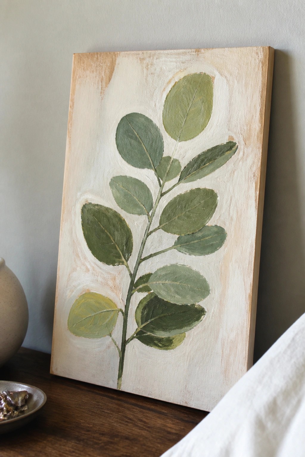 Green leafy branch painting on canvas beside vase on wooden table.
