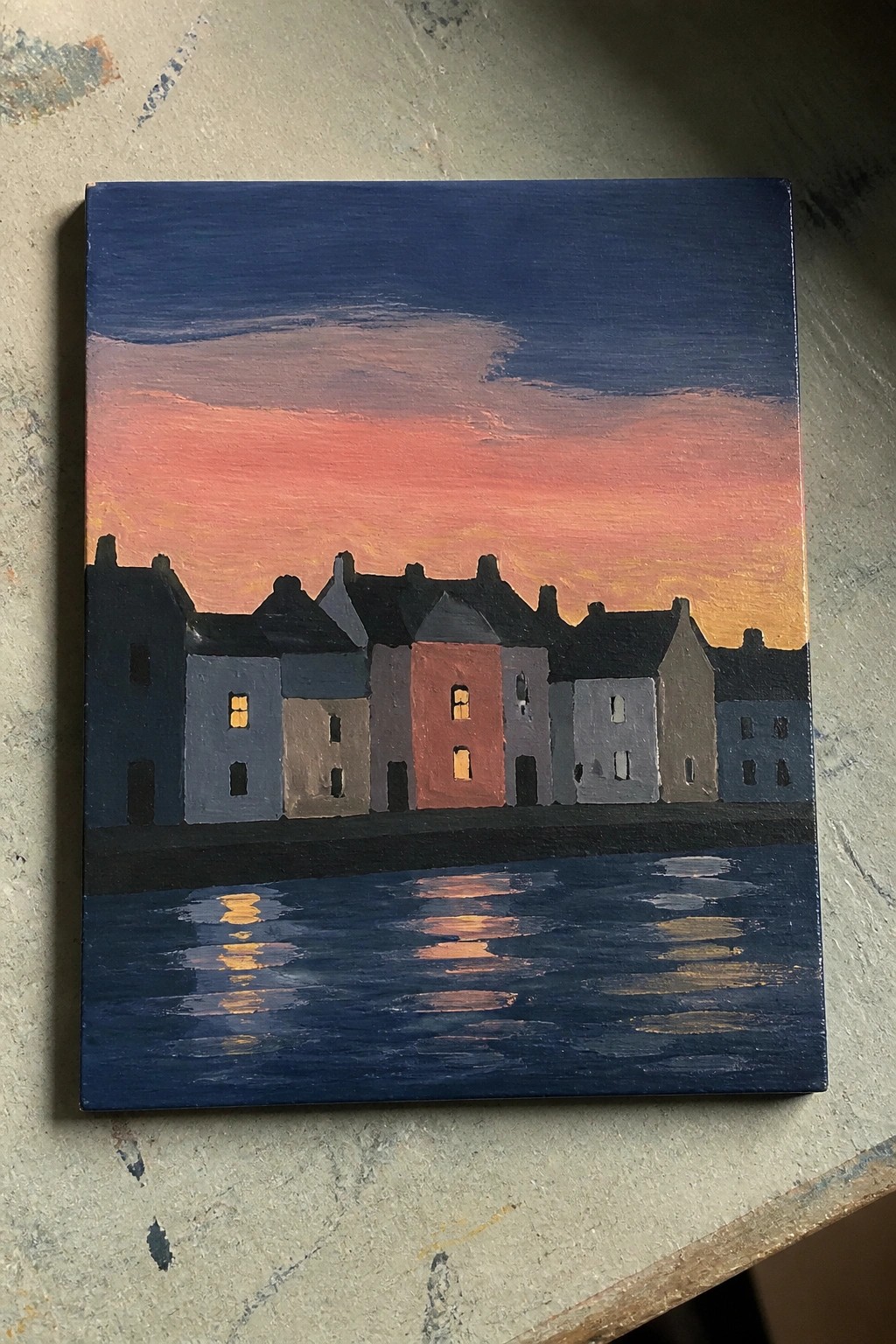 Painted row of houses at dusk with glowing windows reflected in dark blue water under pink sky.