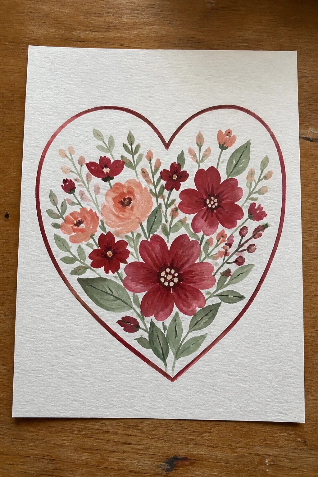 A painting of red flowers and green leaves arranged inside a red heart outline on white paper.