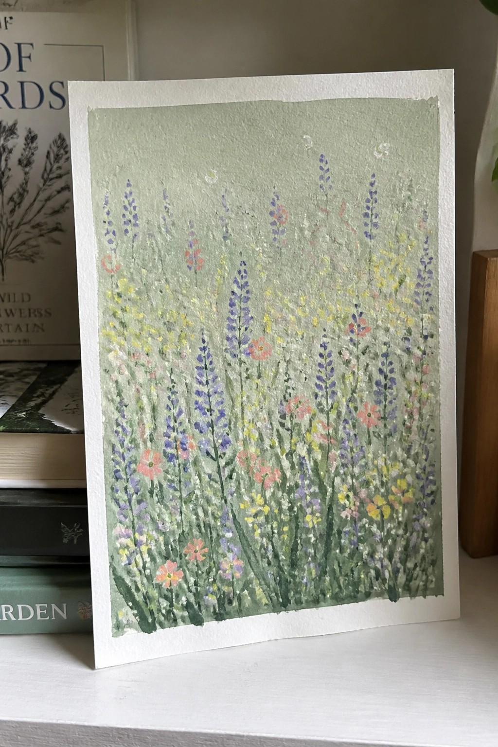 A painting of tall wildflowers in purple, pink, and yellow on a soft green background, displayed on white paper with books in the background.