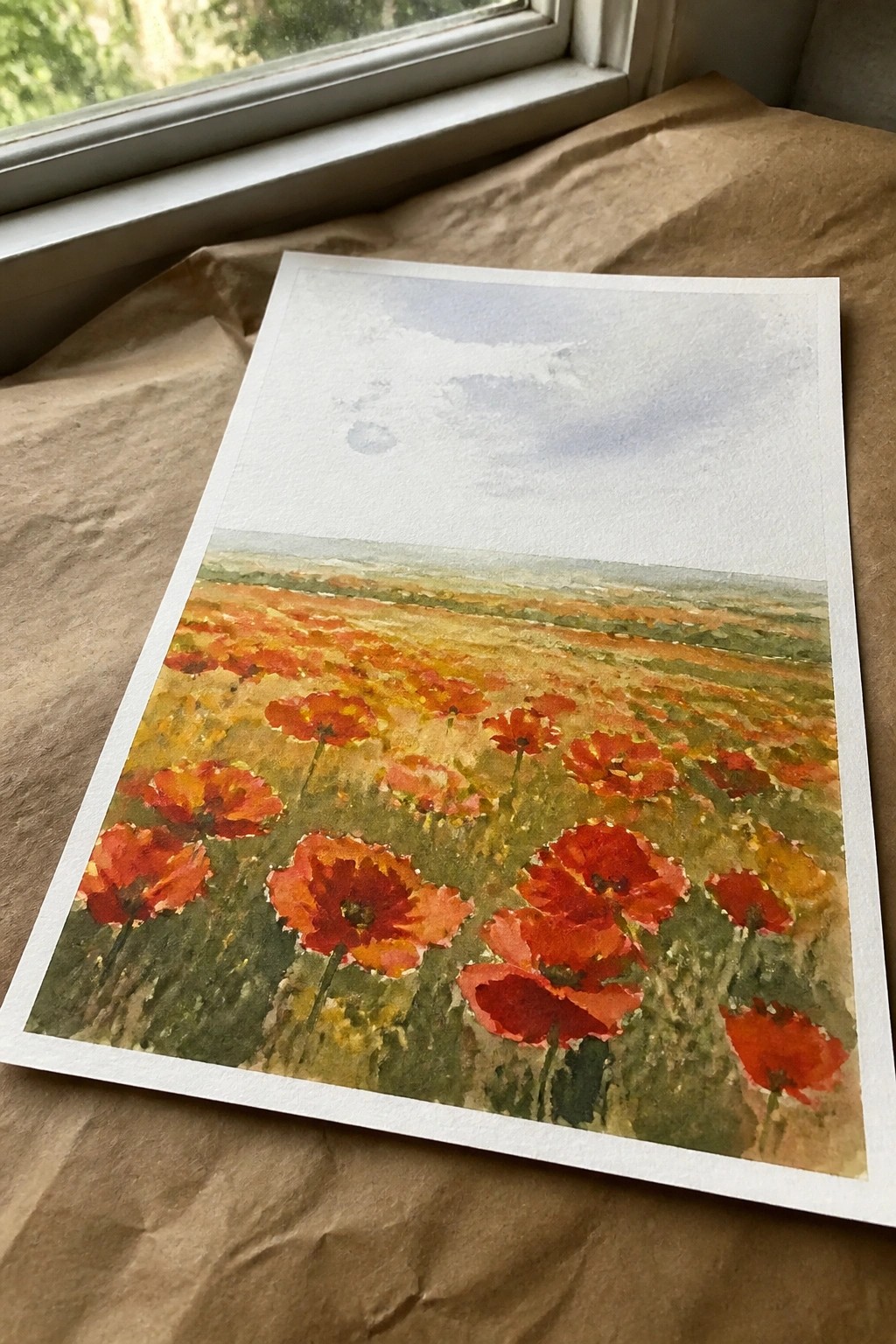 A horizontal painting of red poppies in a field with a pale sky above, placed on brown paper near a window.