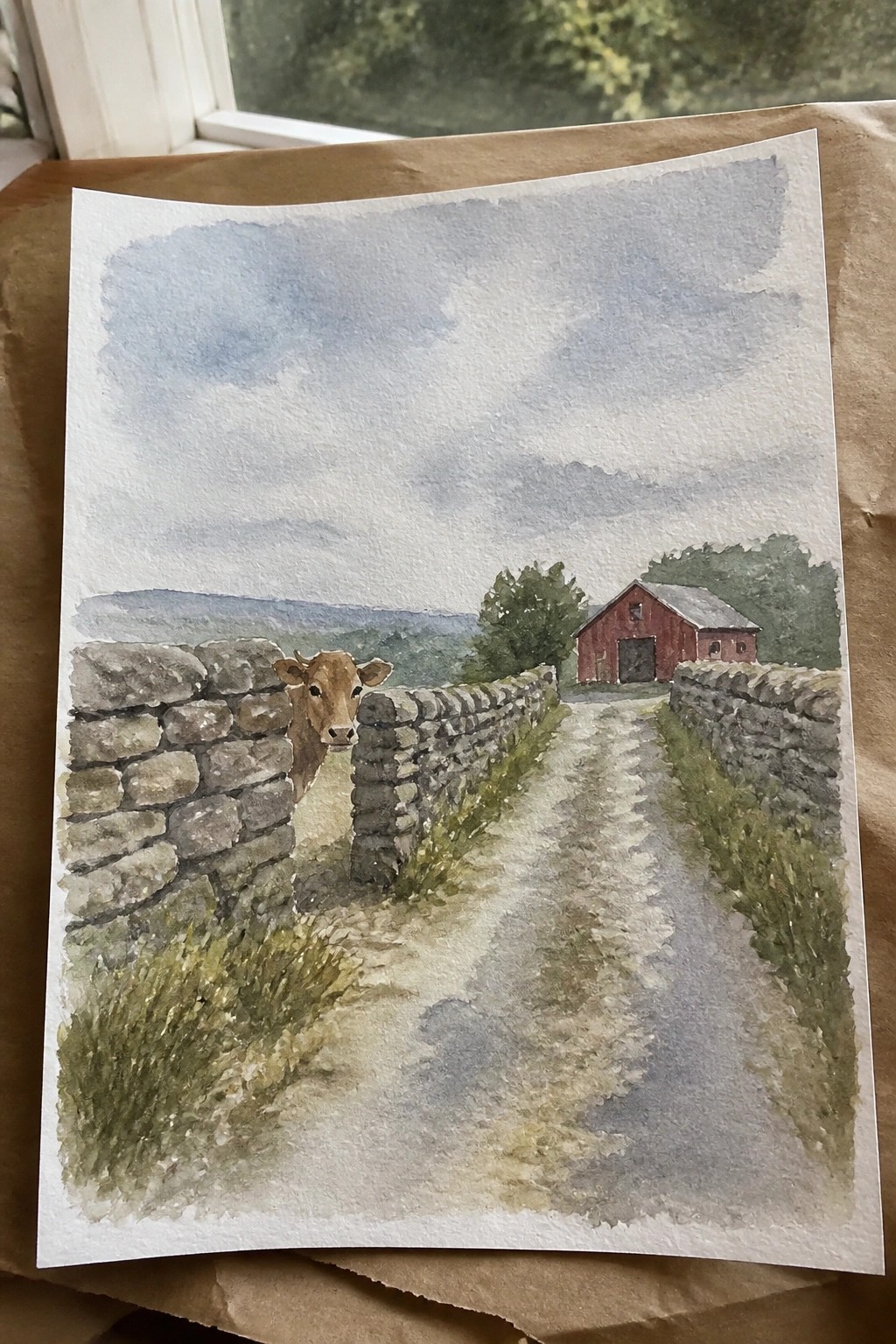 Watercolor of cow peeking over stone wall along path to red barn under clouds