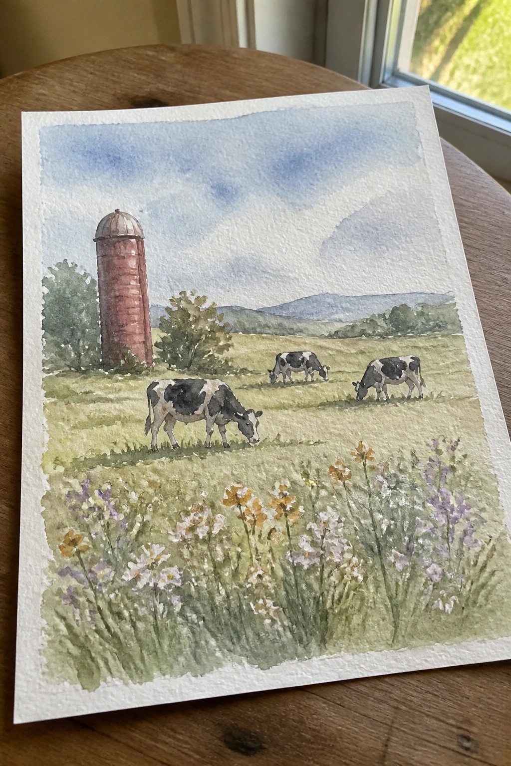 Watercolor of three spotted cows grazing near red silo in wildflower meadow with hills.