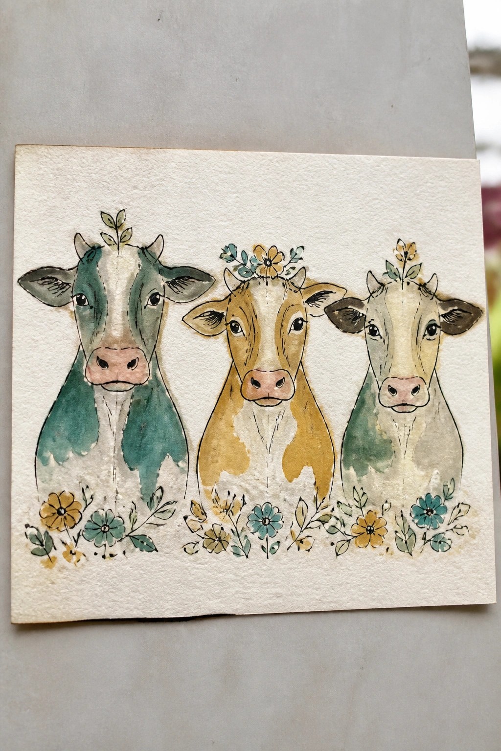 Watercolor painting of three cows with floral crowns above decorative flowers.