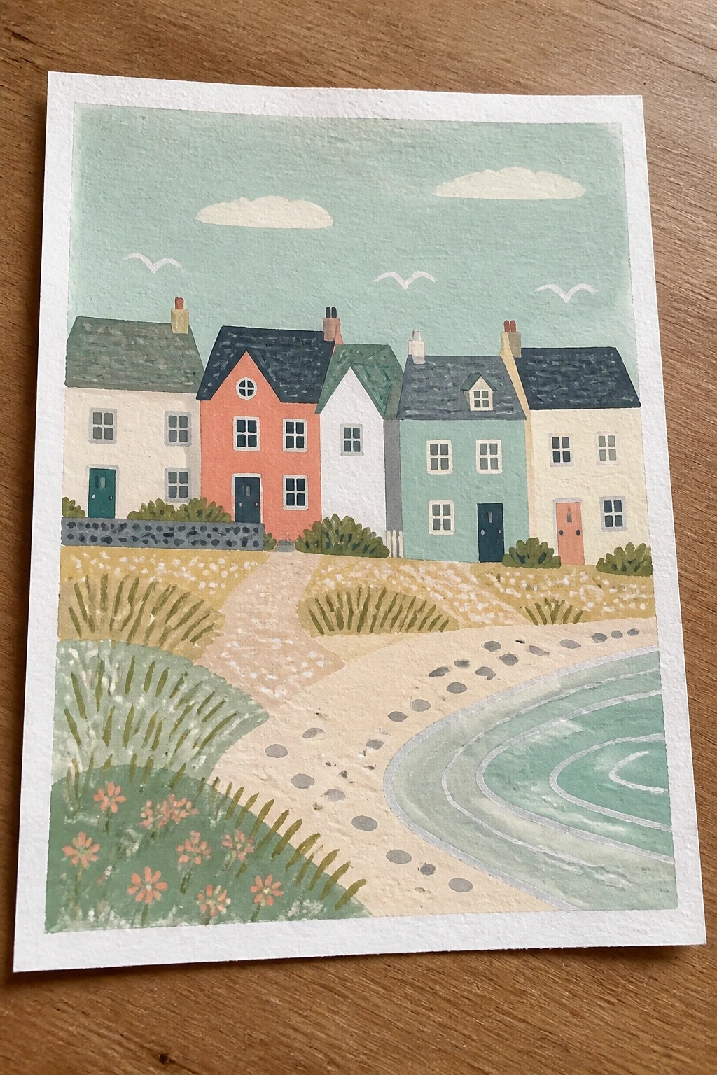 Colorful seaside houses line a winding sandy path with flowers and sea views.