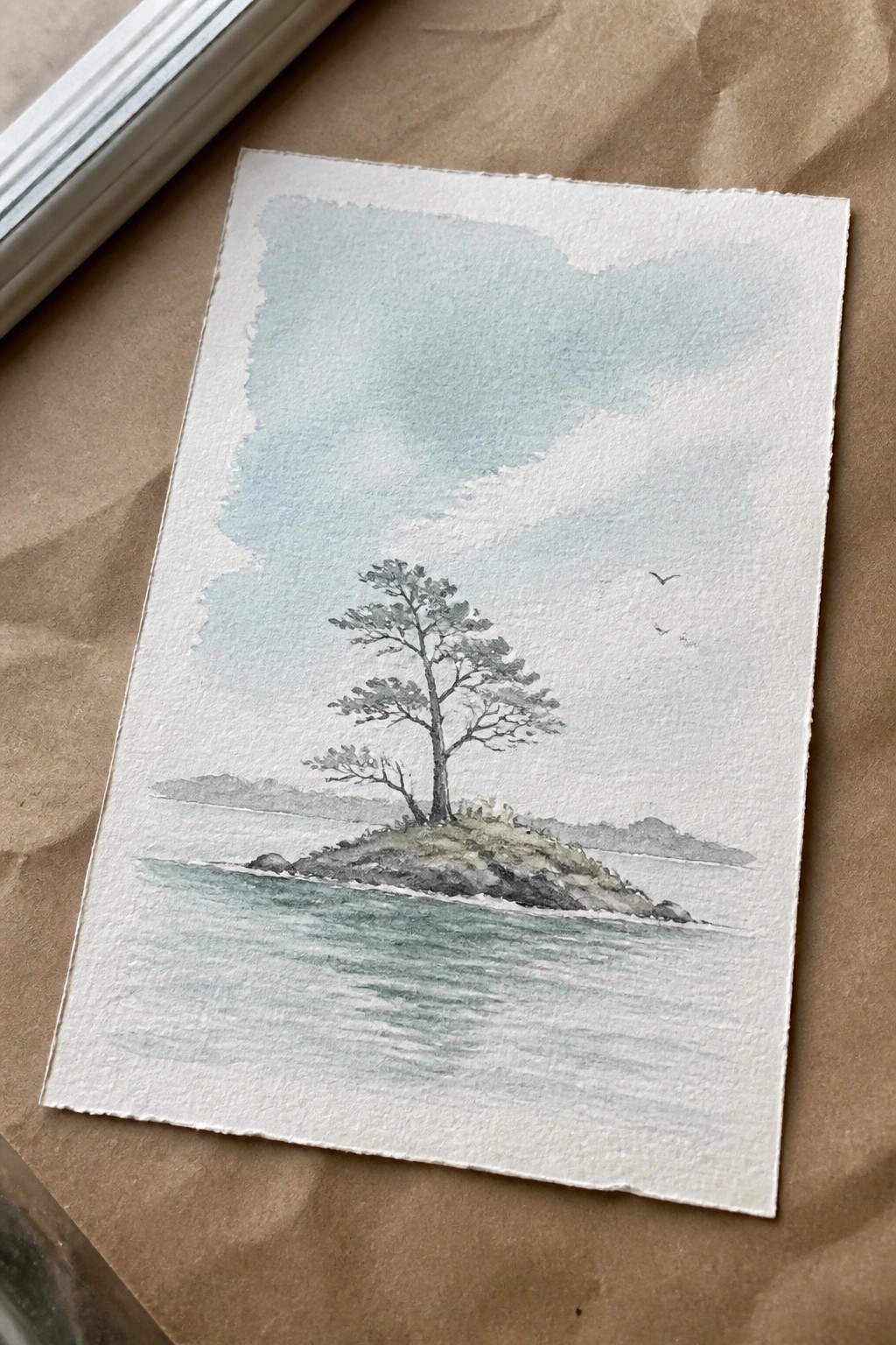 Watercolor of lone pine tree on rocky islet with distant hills, birds, and calm water.