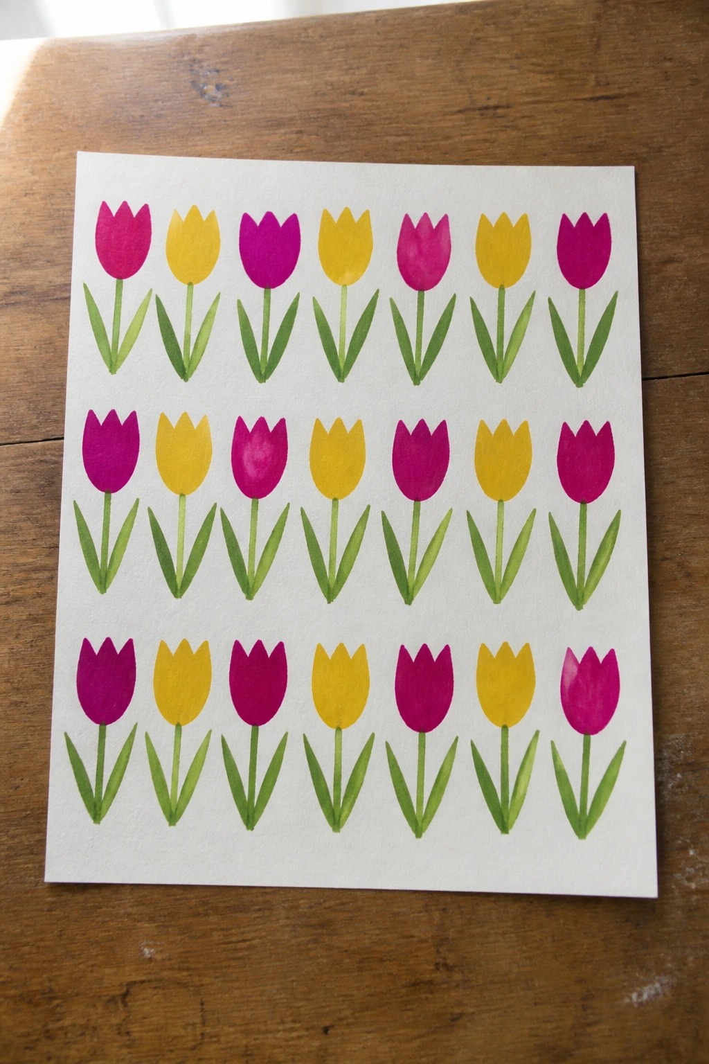 Grid of alternating pink and yellow tulips watercolor painted on white paper on wood.