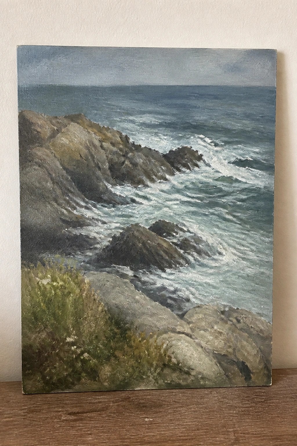 Seascape painting of waves crashing on rocky cliffs under gray skies with foreground plants