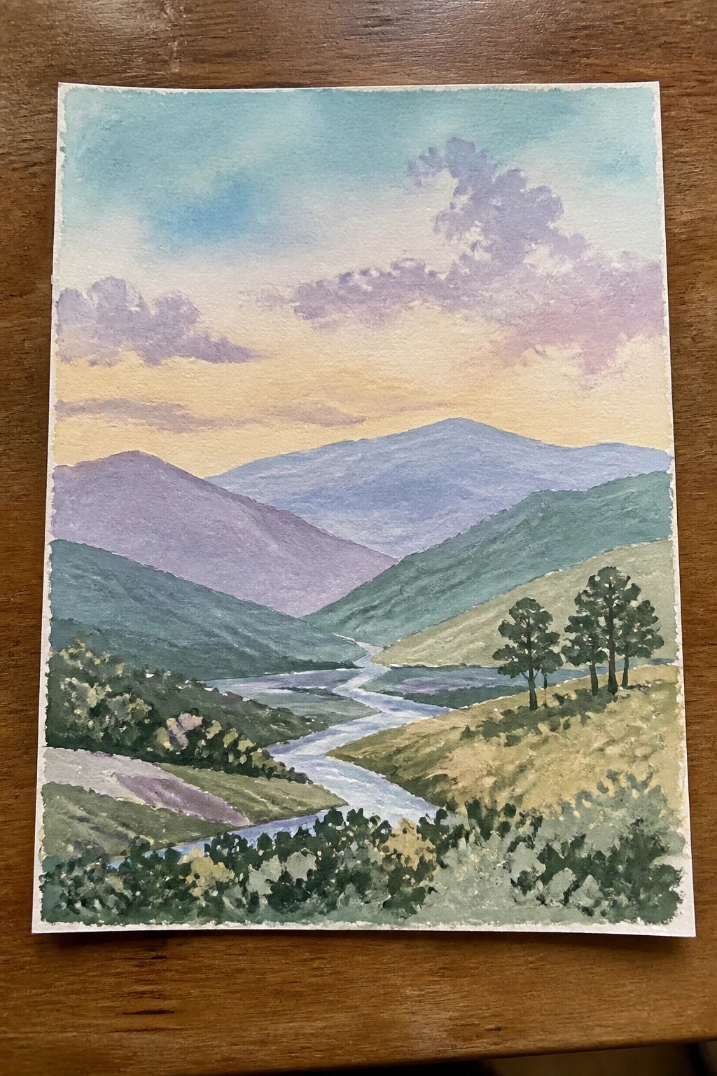 Watercolor landscape of winding river valley with layered purple mountains under colorful sunset sky.