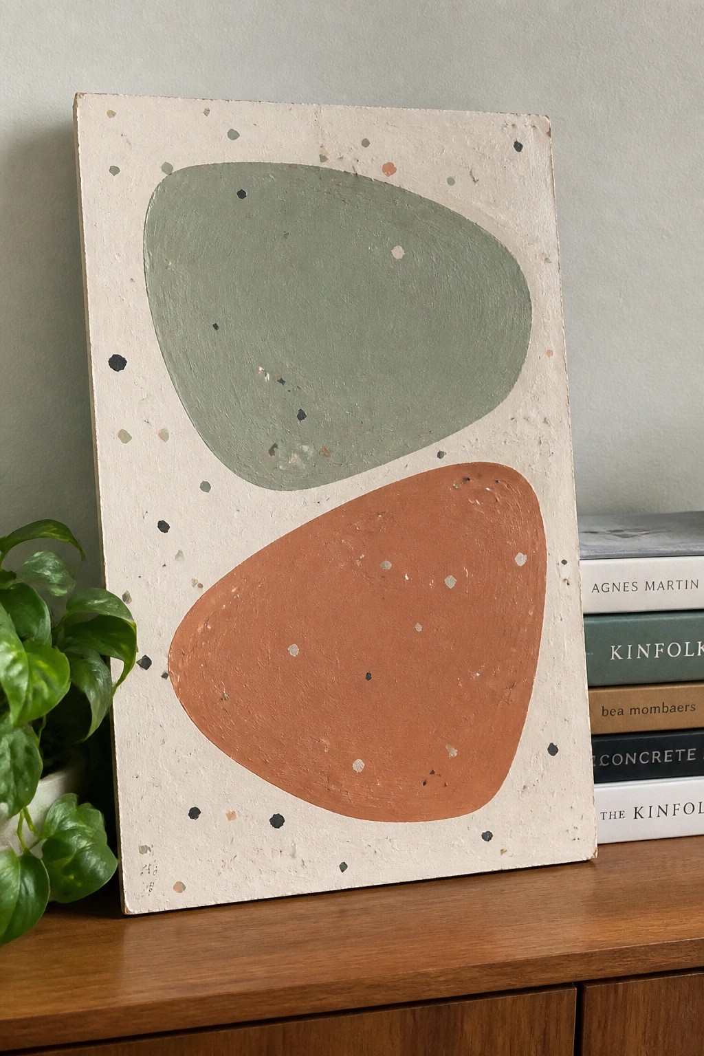 Abstract painting with sage green and terracotta shapes on speckled beige, beside plant and books