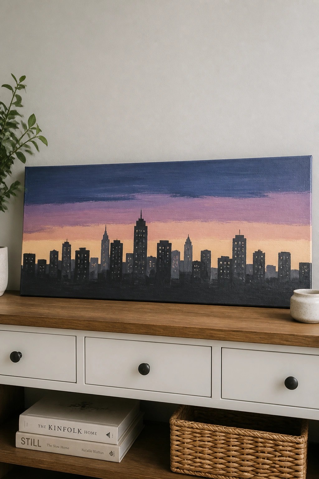 Dark city skyline painting on wooden console with books and woven basket below