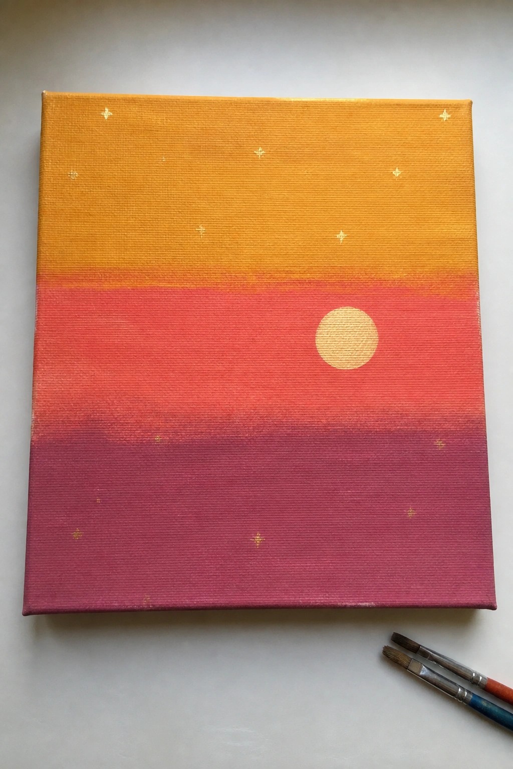 Canvas painting of orange-pink gradient with yellow sun, gold stars, brushes nearby.