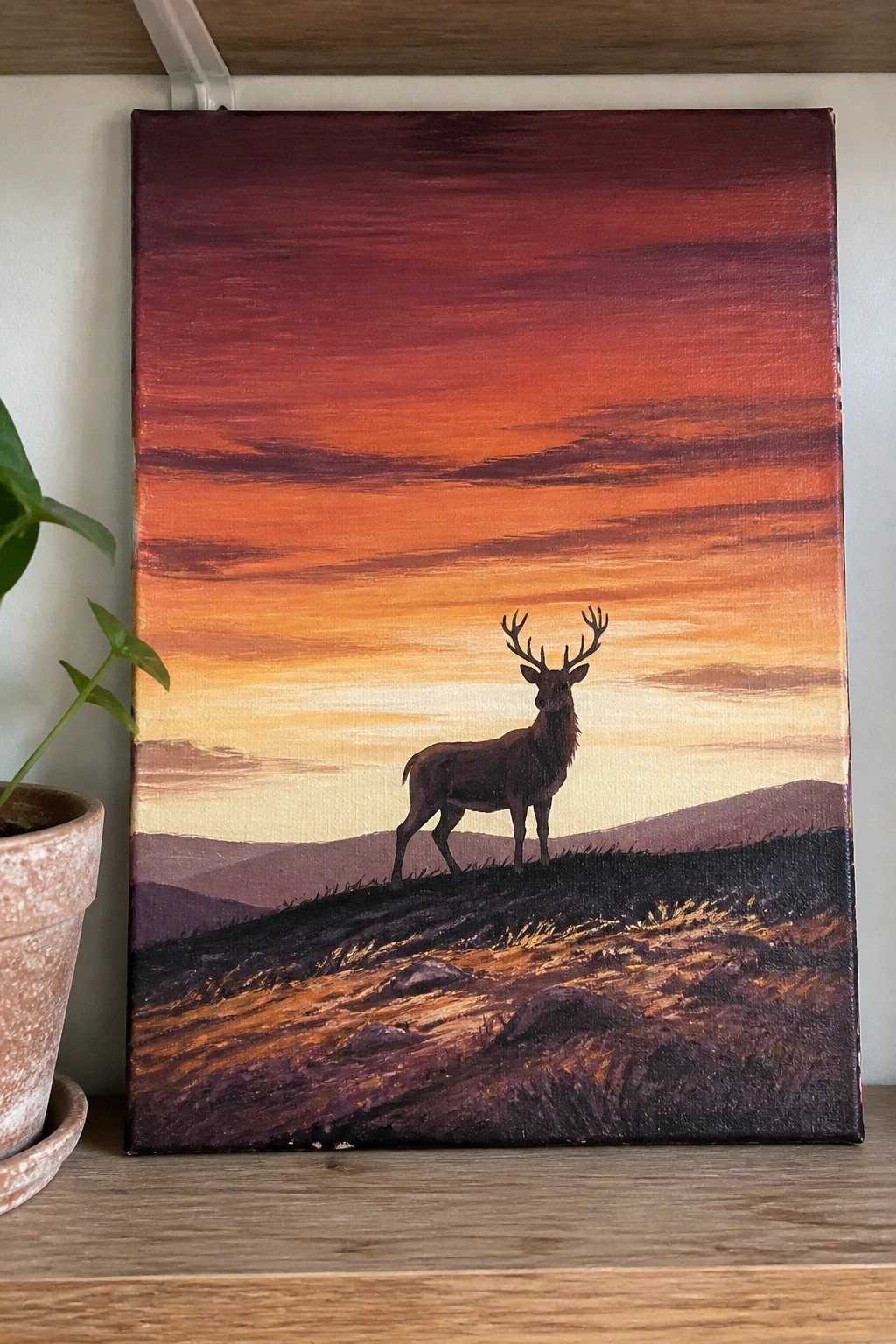Dark stag with antlers on hill under red-orange sunset sky, potted plant beside painting.