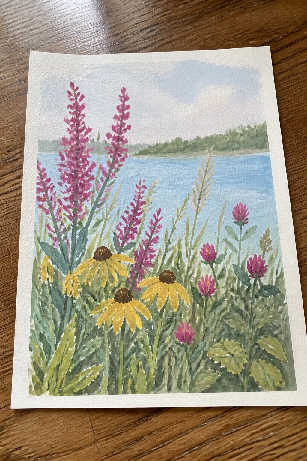 Watercolor wildflowers with purple spikes, yellow blooms, and pink clover by blue water