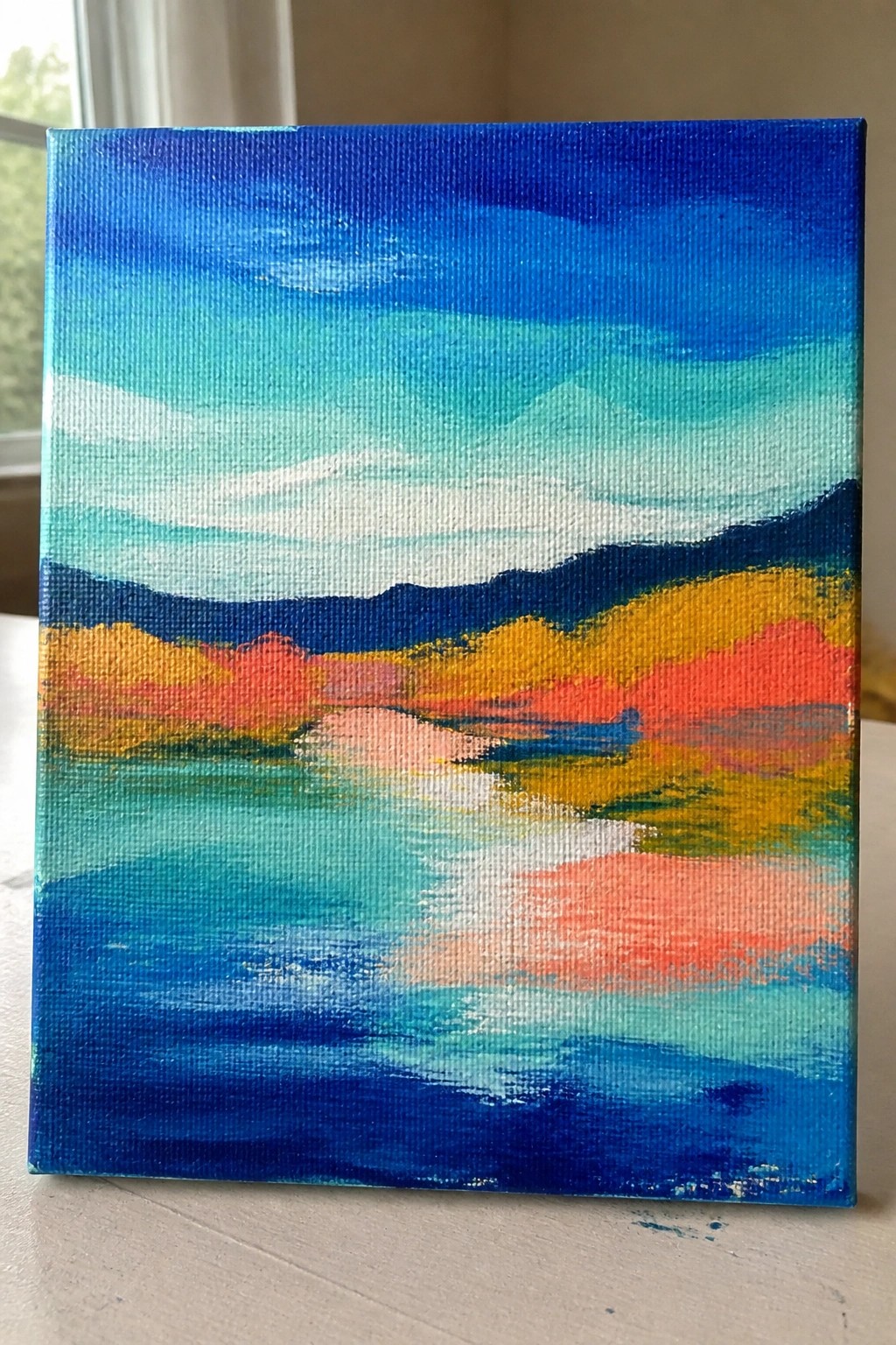 Abstract canvas painting of vibrant mountains and water in bold blues, yellows, and reds.