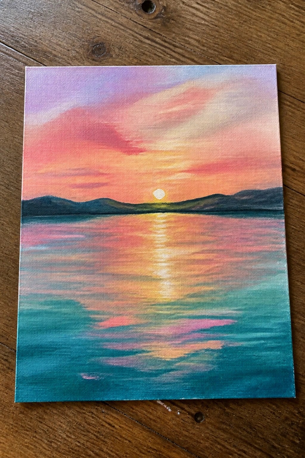 Colorful sunset painting with sun reflecting on water below mountain silhouette.