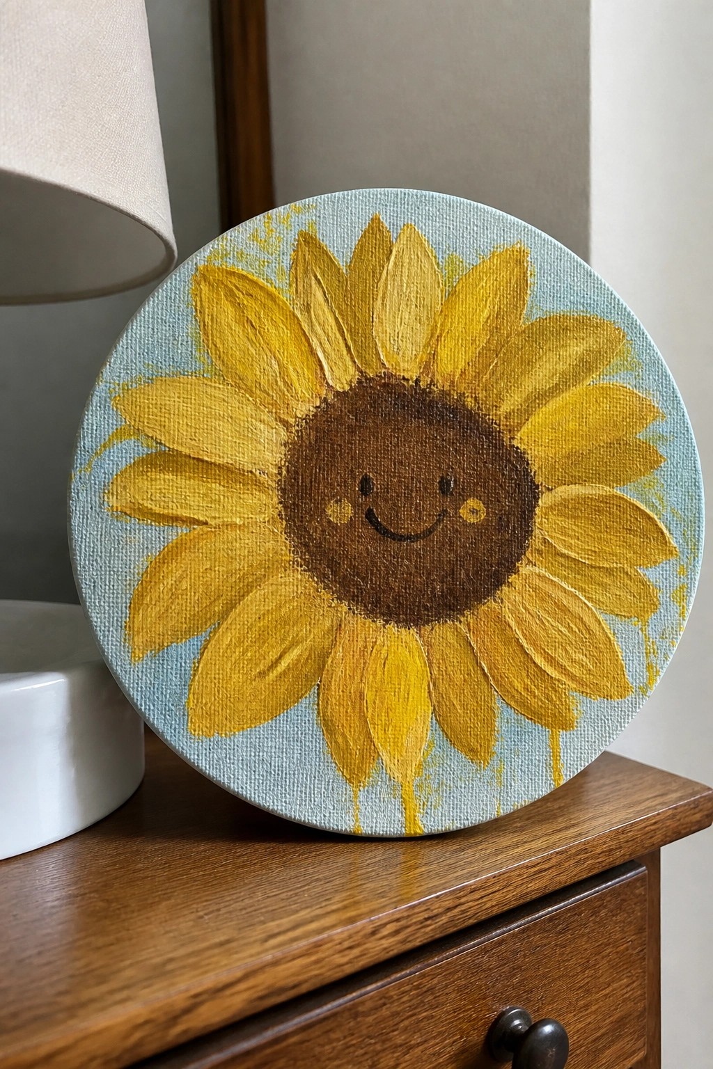 Round canvas painting of smiling sunflower with yellow petals on wooden surface.