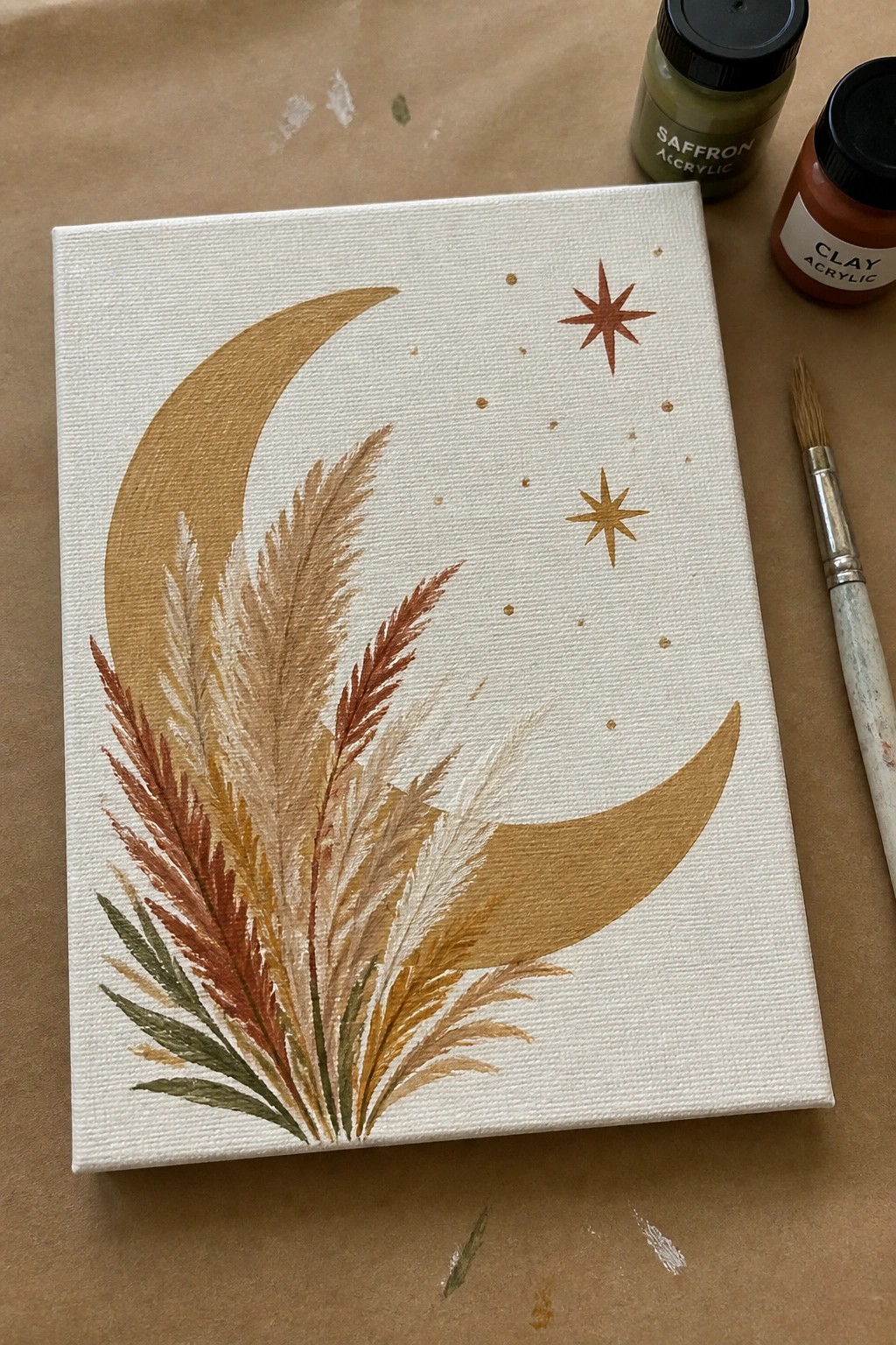 Canvas painting of golden crescent moon, stars and pampas grass beside acrylic paints and brush