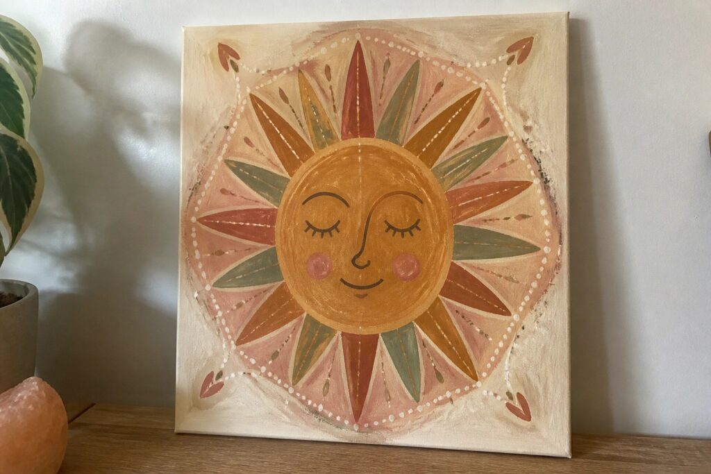 A canvas painting of a smiling sun face with closed eyes in warm orange and yellow tones, surrounded by pointed rays and topped and bottomed with green leaf garlands on a beige background.