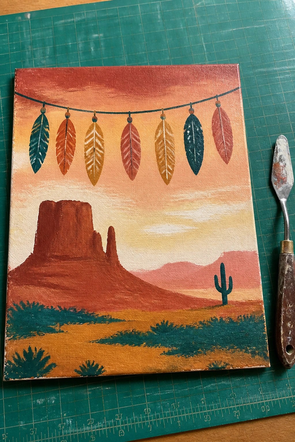 A square acrylic painting of a desert landscape at sunset with mesas and a cactus, topped by a string of colorful feathers.