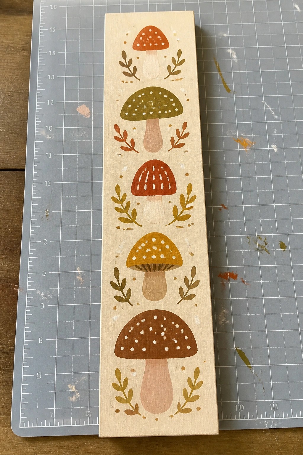 Vertical wooden plaque with five stacked colorful mushrooms and leafy accents on cutting mat.