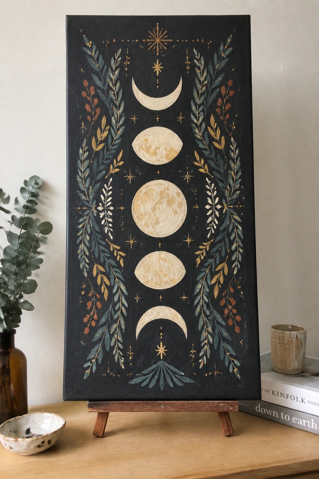 Moon phase painting with teal leaves and gold stars on black canvas easel