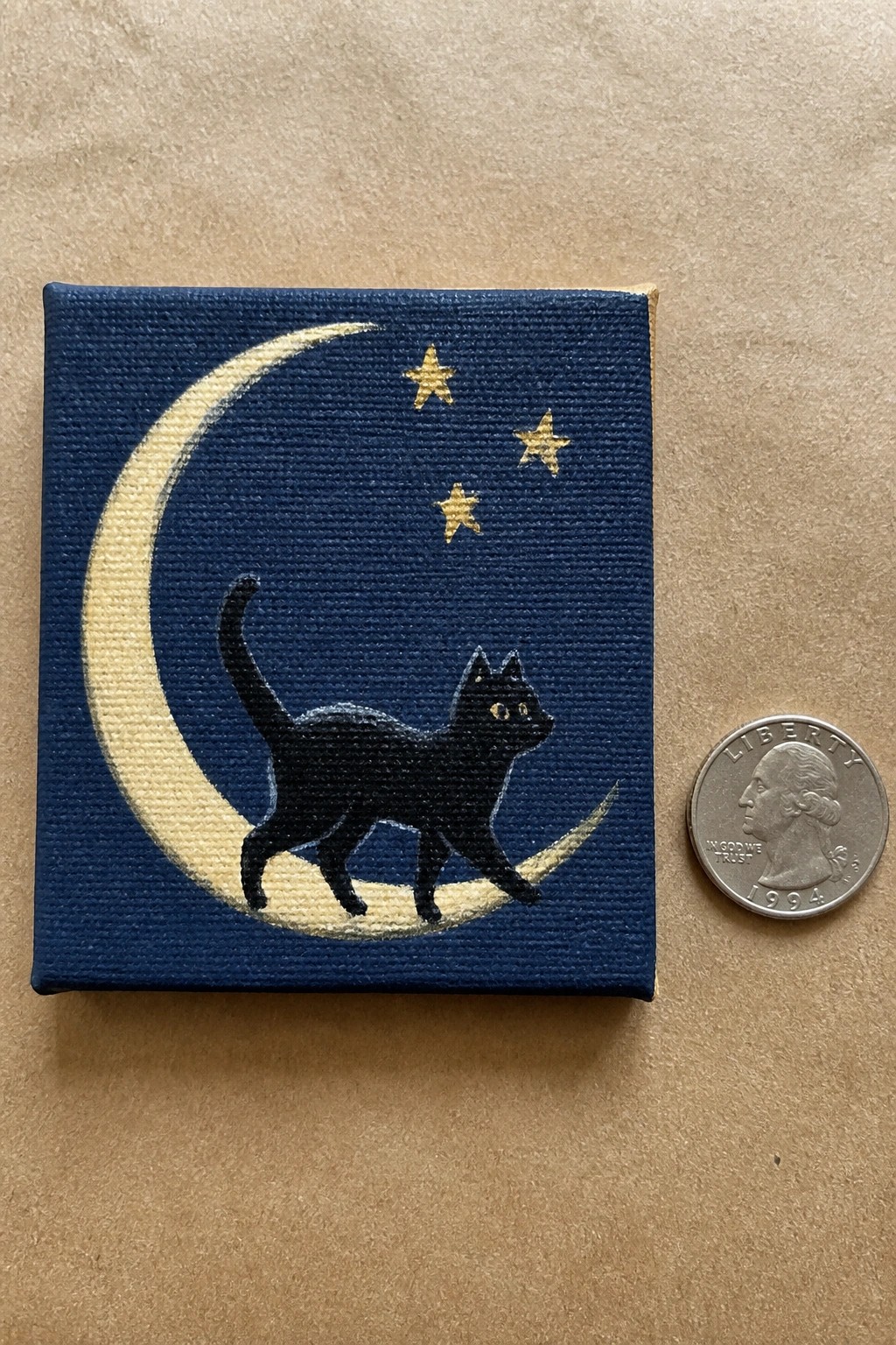 Black cat on yellow crescent moon with gold stars on blue canvas beside a quarter.