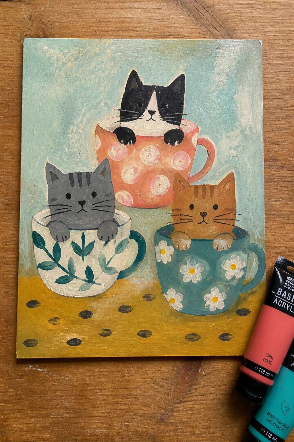 Three cats peeking from patterned mugs on yellow polka-dotted surface in acrylic painting