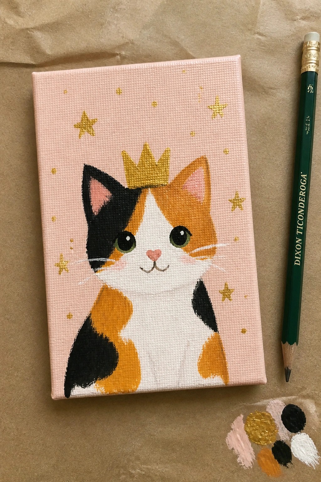 Crowned calico cat painting on pink starry canvas beside green pencil and paint dabs