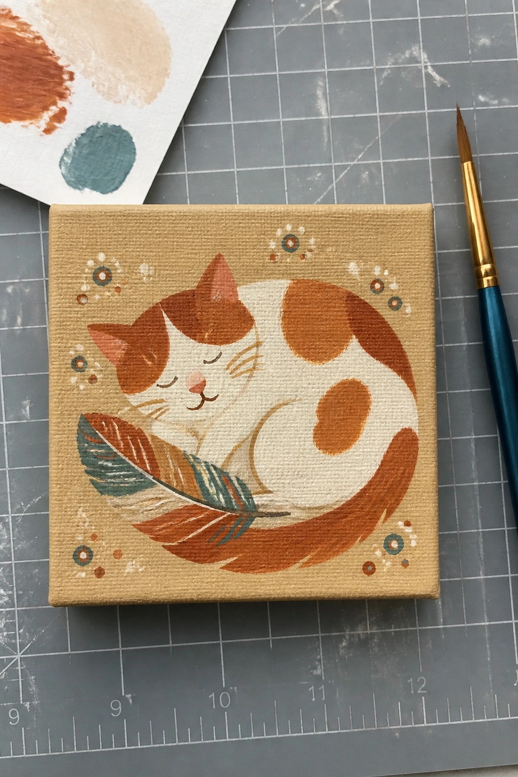 Sleeping calico cat curled with feather on tan canvas beside paintbrush on grid mat.