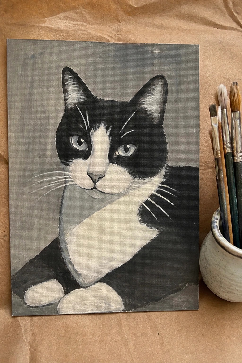 Black and white cat painting on canvas beside paintbrushes in ceramic holder
