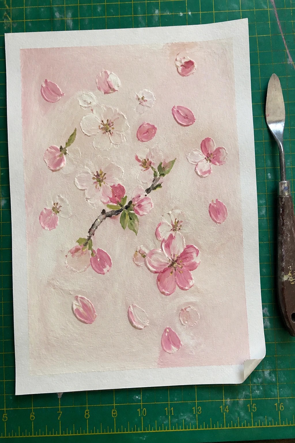Pink cherry blossom painting with scattered petals beside palette knife on green mat.