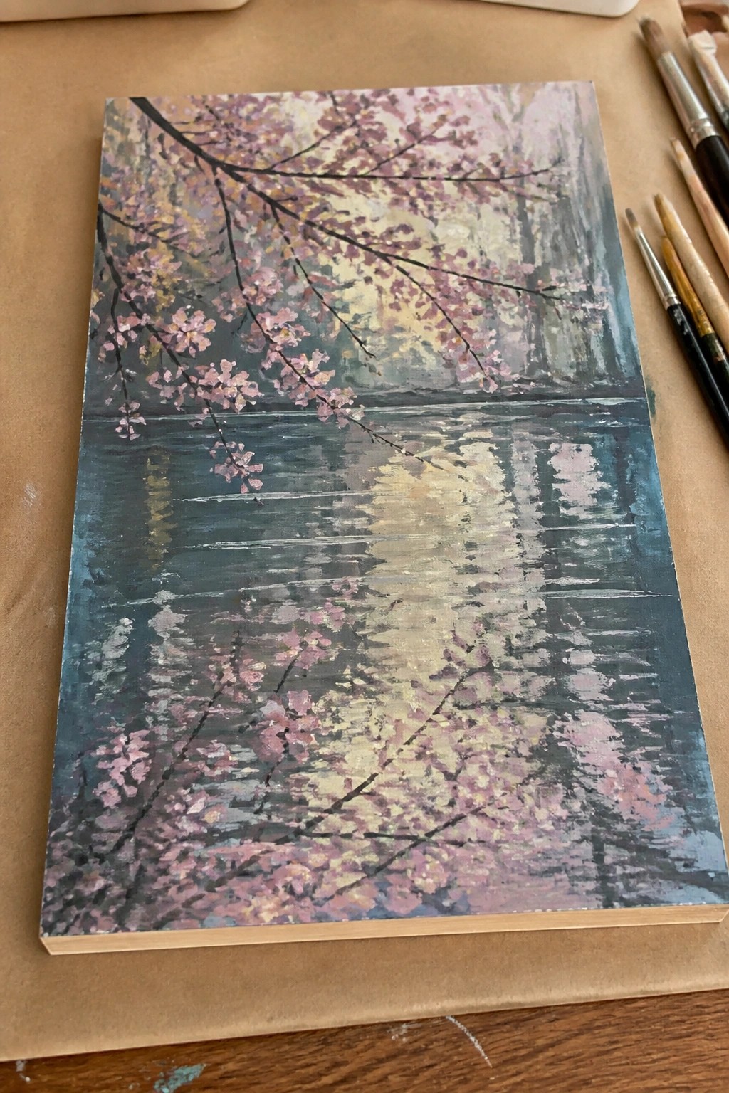 Impressionistic painting of pink cherry blossoms reflected in blue water beside paintbrushes.