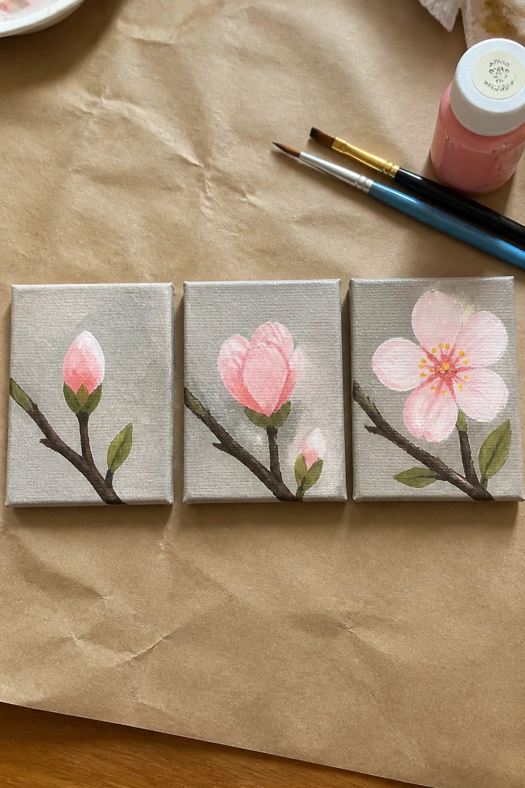 Three small canvases with pink cherry blossoms from bud to full bloom beside paint supplies