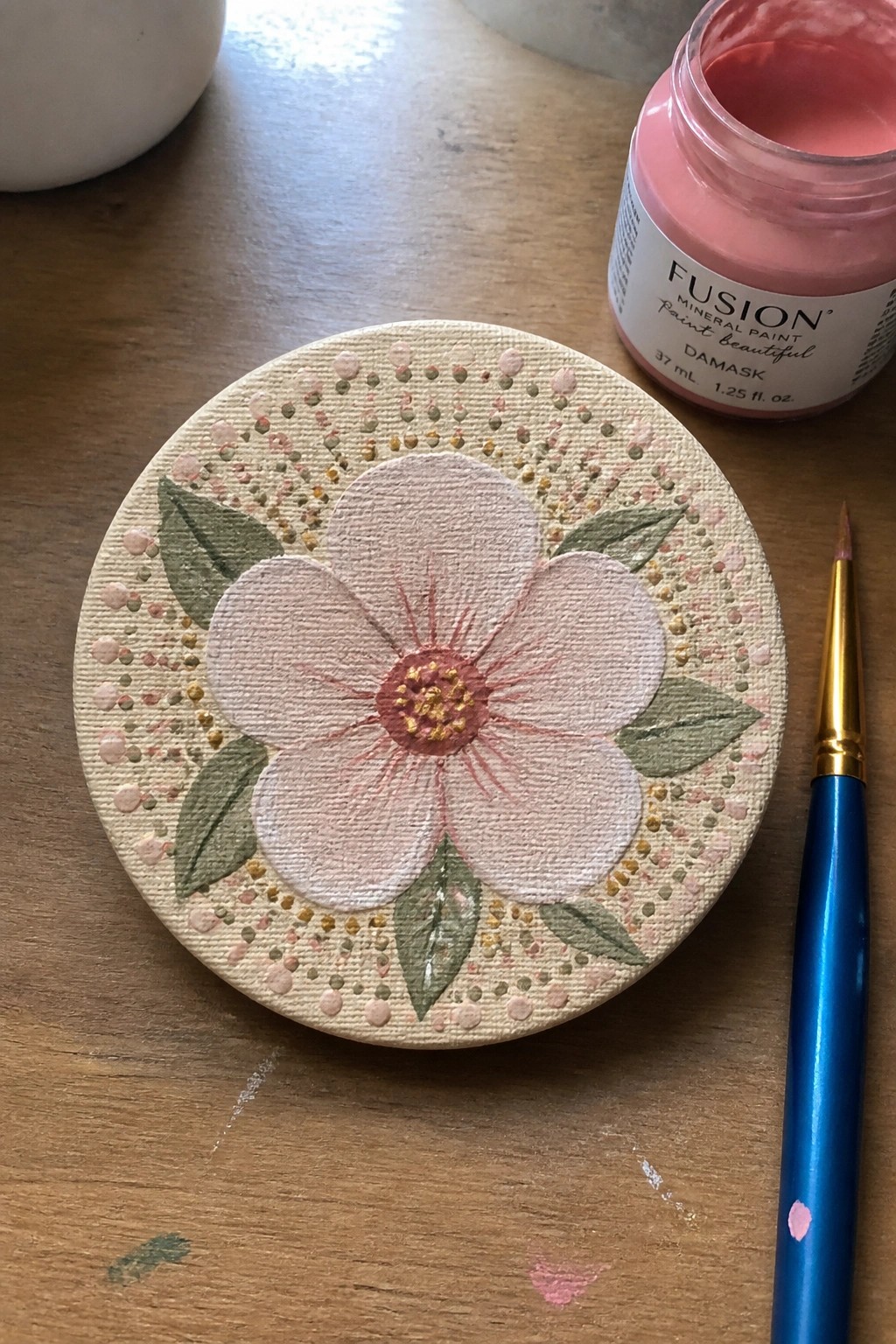 Circular canvas with pink flower, green leaves, dotted border beside paint jar and brush