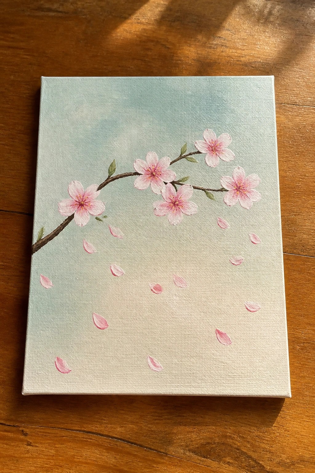 Pink cherry blossom branch with falling petals on light blue canvas over wood