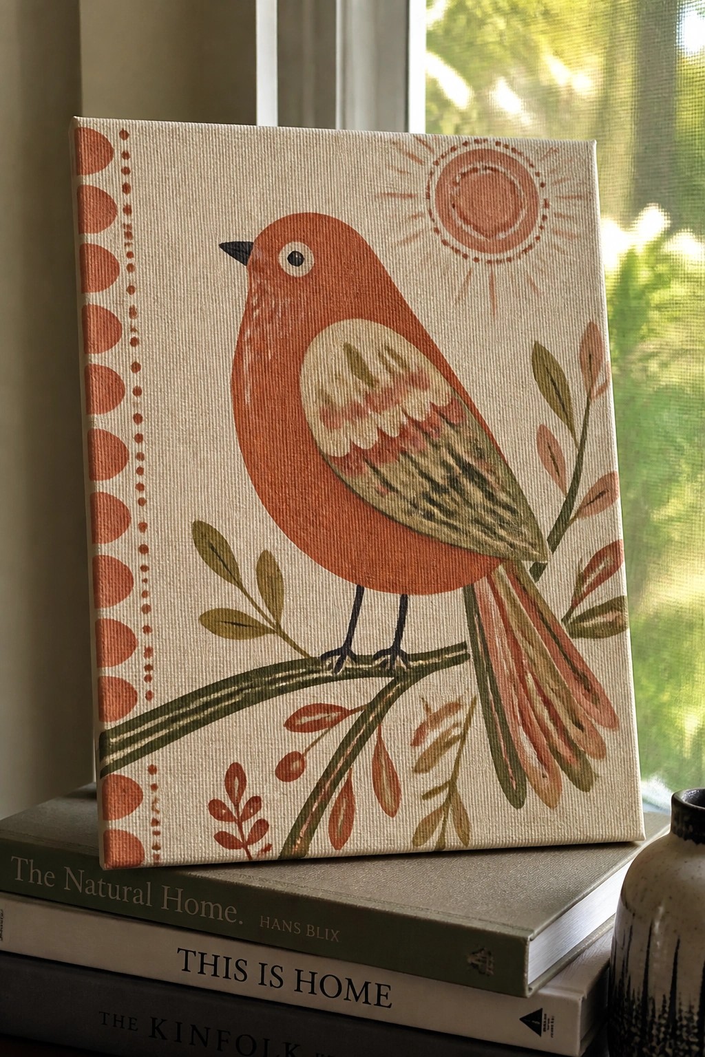 Orange-red bird on branch with sun, canvas atop stacked books near window