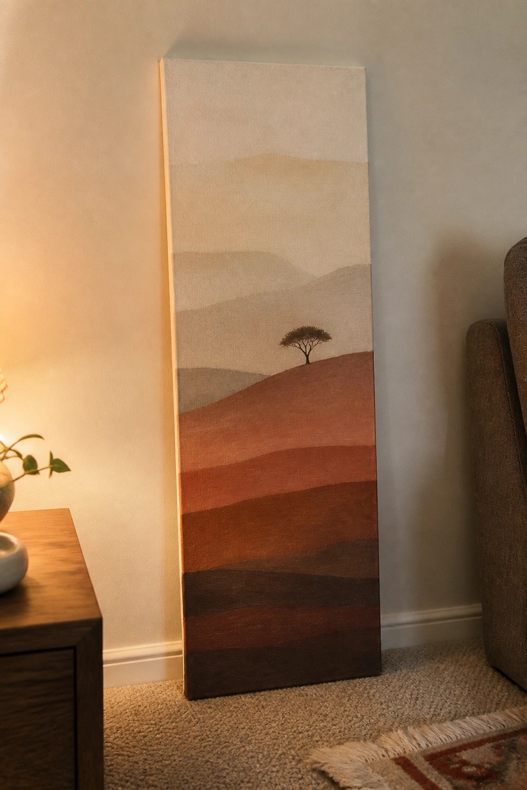 Tall vertical canvas painting of layered earth-tone hills with a lone tree in a room