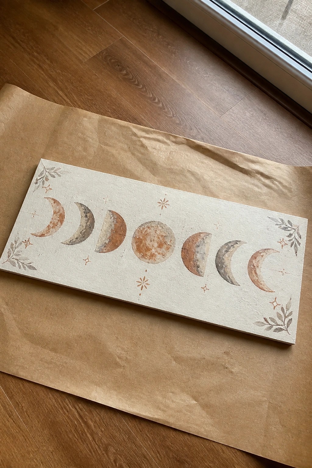 Moon phase artwork with stars and leaves on beige board over brown paper.